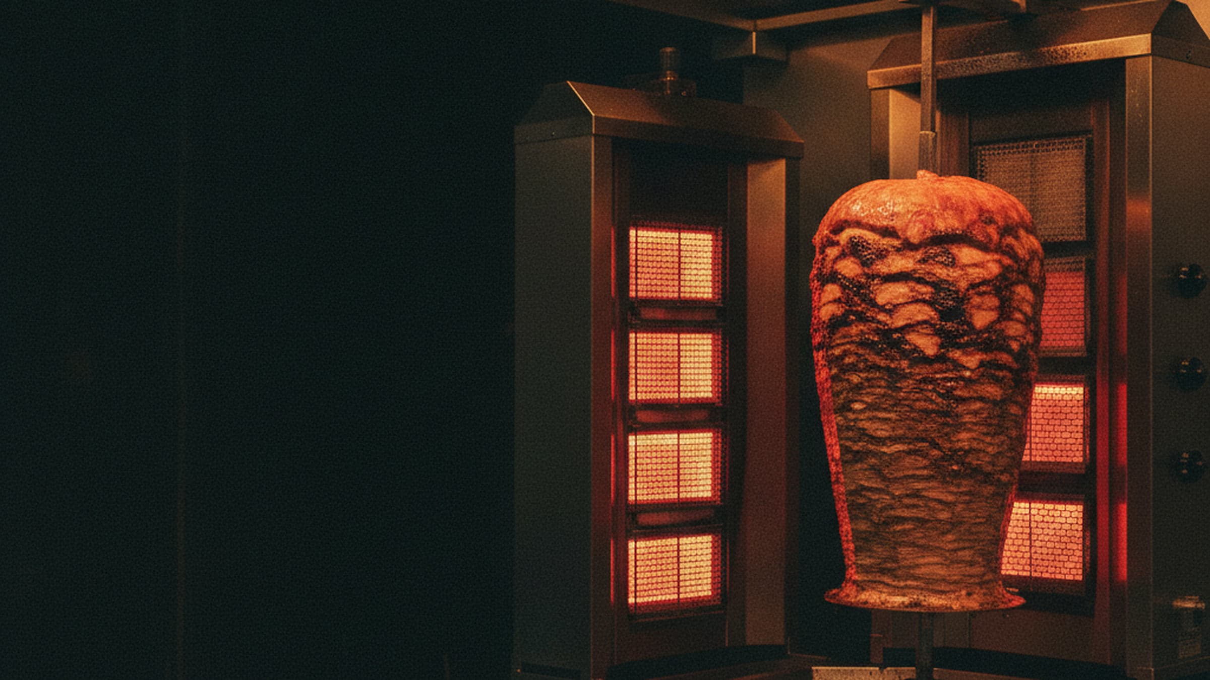 Ein Dönerspieß dreht sich vor den glühenden Heizstäben eines Grills in einer dunklen Imbissküche